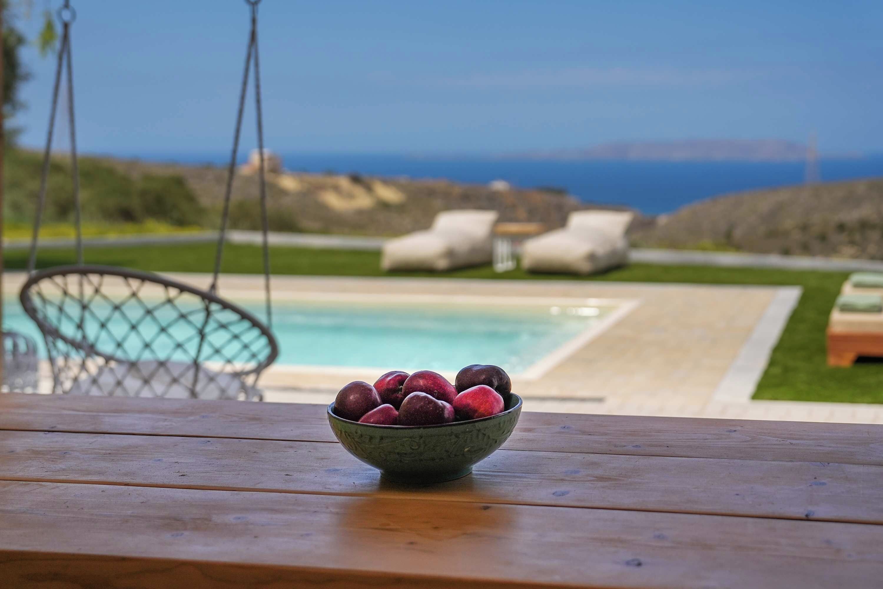 A wooden table with a bowl of fruit in the foreground, a pool, and lounge chairs in the background.
