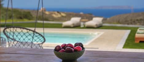 A wooden table with a bowl of fruit in the foreground, a pool, and lounge chairs in the background.
