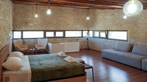 Ein geräumiges Zimmer mit einem großen Bett, einer Couch und einer Sitzecke. Mehrere Fenster sorgen für natürliches Licht und eine Steinwand ist zu sehen.