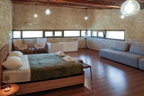 Ein geräumiges Zimmer mit einem großen Bett, einer Couch und einer Sitzecke. Mehrere Fenster sorgen für natürliches Licht und eine Steinwand ist zu sehen.
