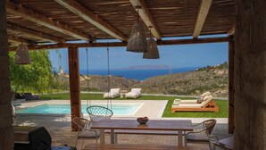 Ein überdachter Außenbereich mit Holztisch und Stühlen, einem Grill und Blick auf Pool und Landschaft.