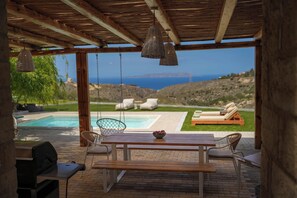 Ein überdachter Außenbereich mit Holztisch und Stühlen, einem Grill und Blick auf Pool und Landschaft.