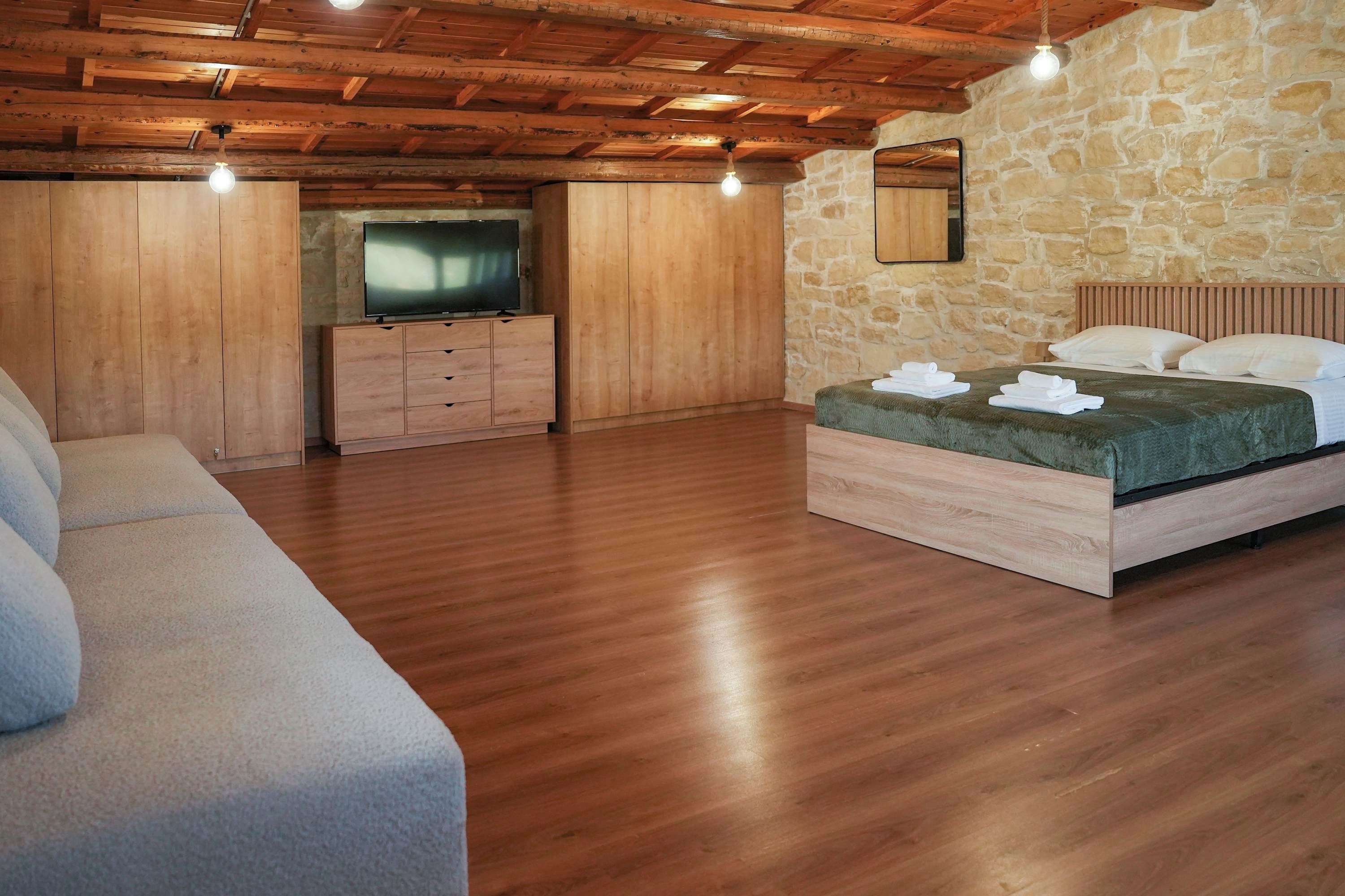 A bedroom with a wooden ceiling, stone walls, a bed with white linens, a TV, and a wooden cabinet.