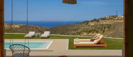 A poolside area with a view of the sea, a wooden table with a bowl of fruit, and two lounge chairs.