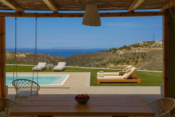 Ein Poolbereich mit Meerblick, ein Holztisch mit einer Obstschale und zwei Liegestühle.
