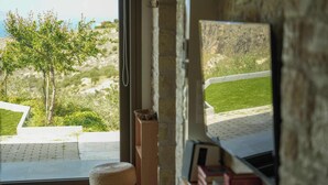 Une pièce avec vue sur un espace verdoyant et un arbre. On y trouve une fenêtre, un tabouret en bois et un mur en pierre.