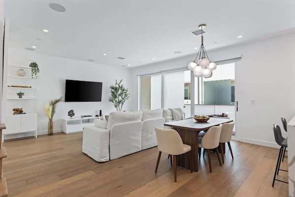A modern living room with a white sofa, a dining table with chairs, a flat-screen TV mounted on the wall, and a large window.