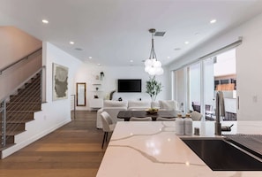 A modern kitchen with a central island, a dining area with a sofa, a staircase, and a large window.