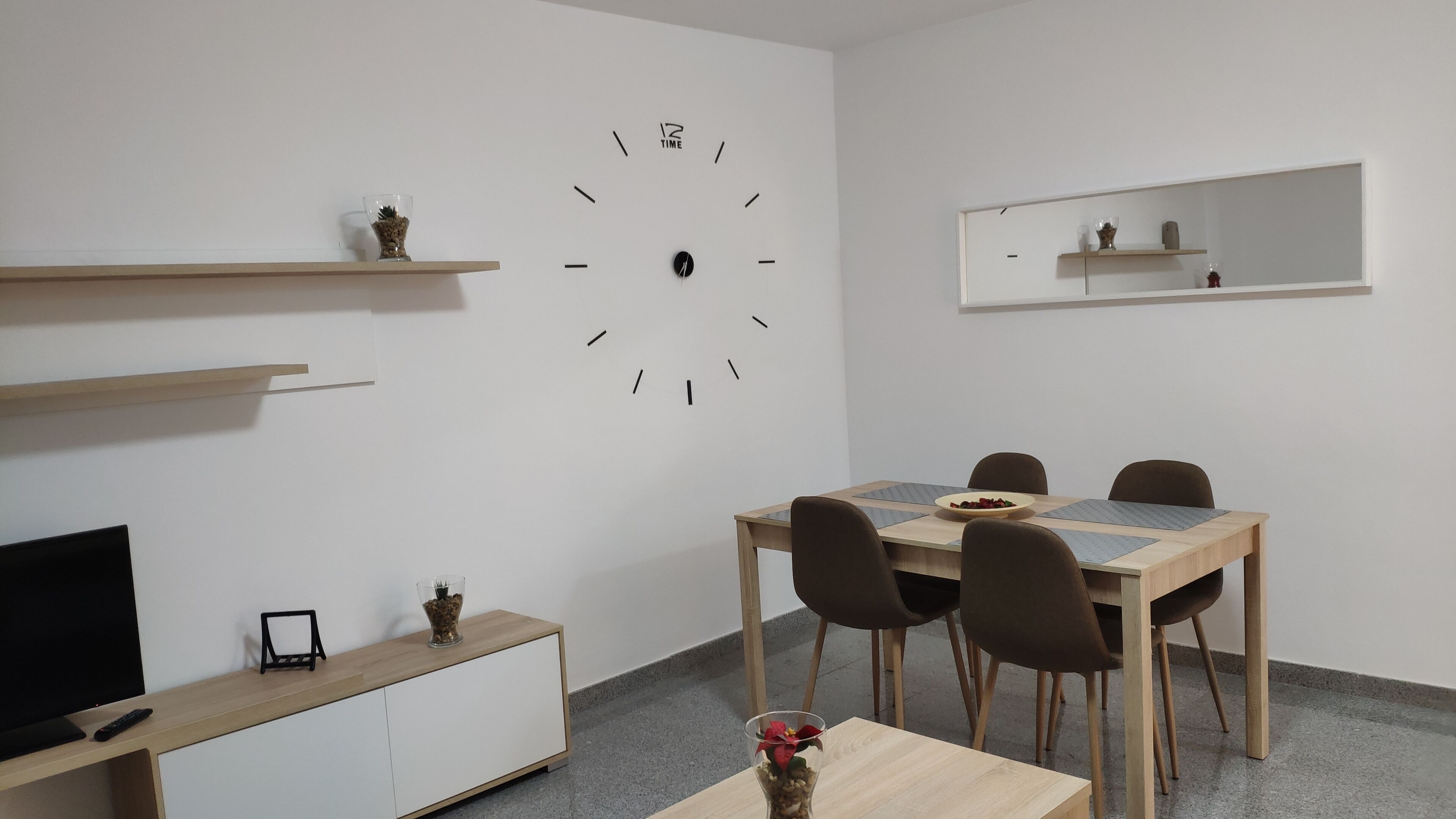 A modern dining area with a wooden table, four chairs, a wall clock, and a television on a cabinet.