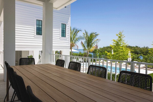 A covered outdoor area with a wooden table and chairs, overlooking a swimming pool and a view of the ocean.