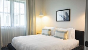 Una cama bien hecha con ropa blanca, un cuadro en la pared y una ventana con cortinas traslúcidas.