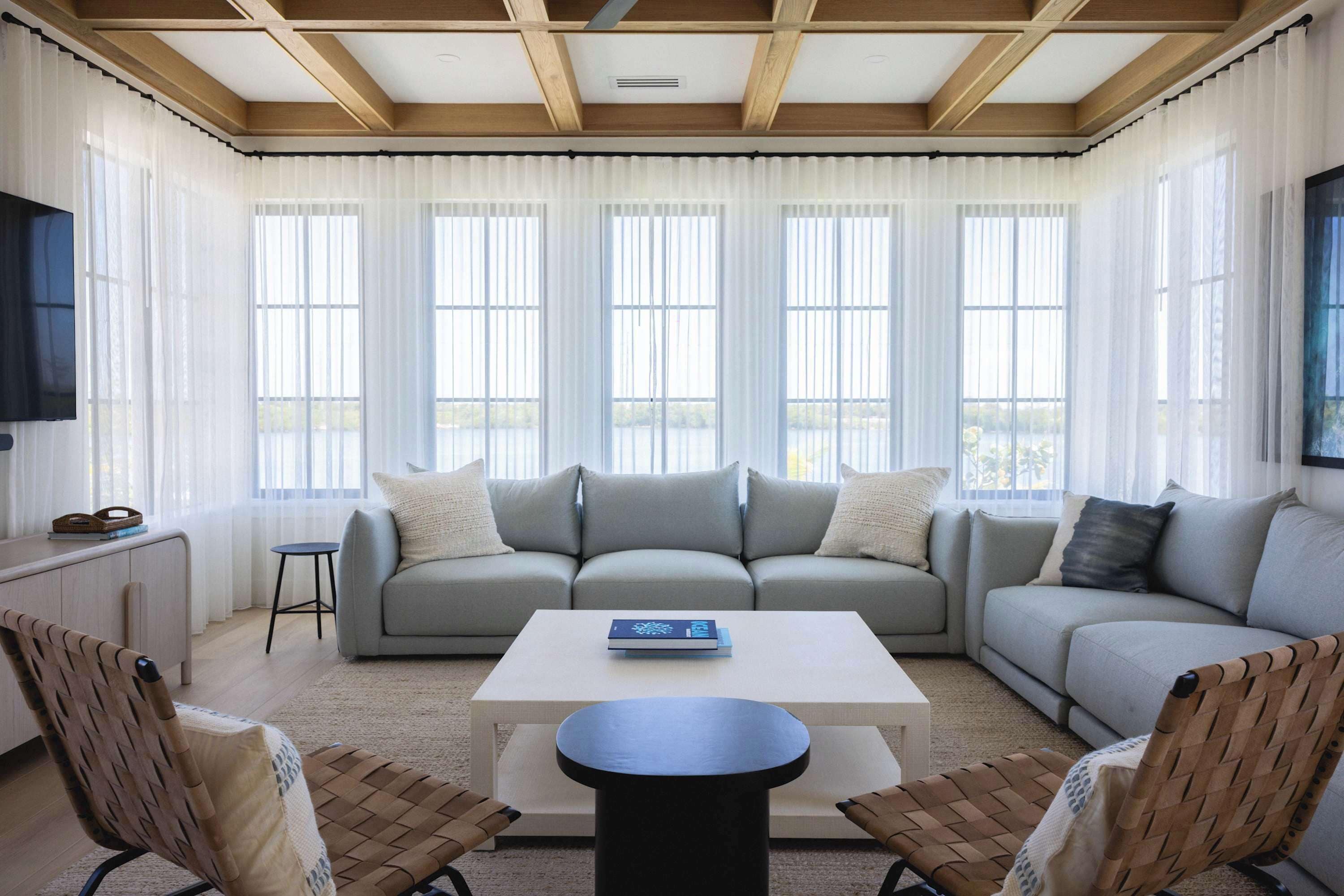 A modern living room with a large sofa, a coffee table, and two chairs. There are large windows with sheer curtains, a flat-screen TV mounted on the wall, and a wooden ceiling.