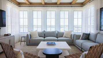 A modern living room with a large sofa, a coffee table, and two chairs. There are large windows with sheer curtains, a flat-screen TV mounted on the wall, and a wooden ceiling.