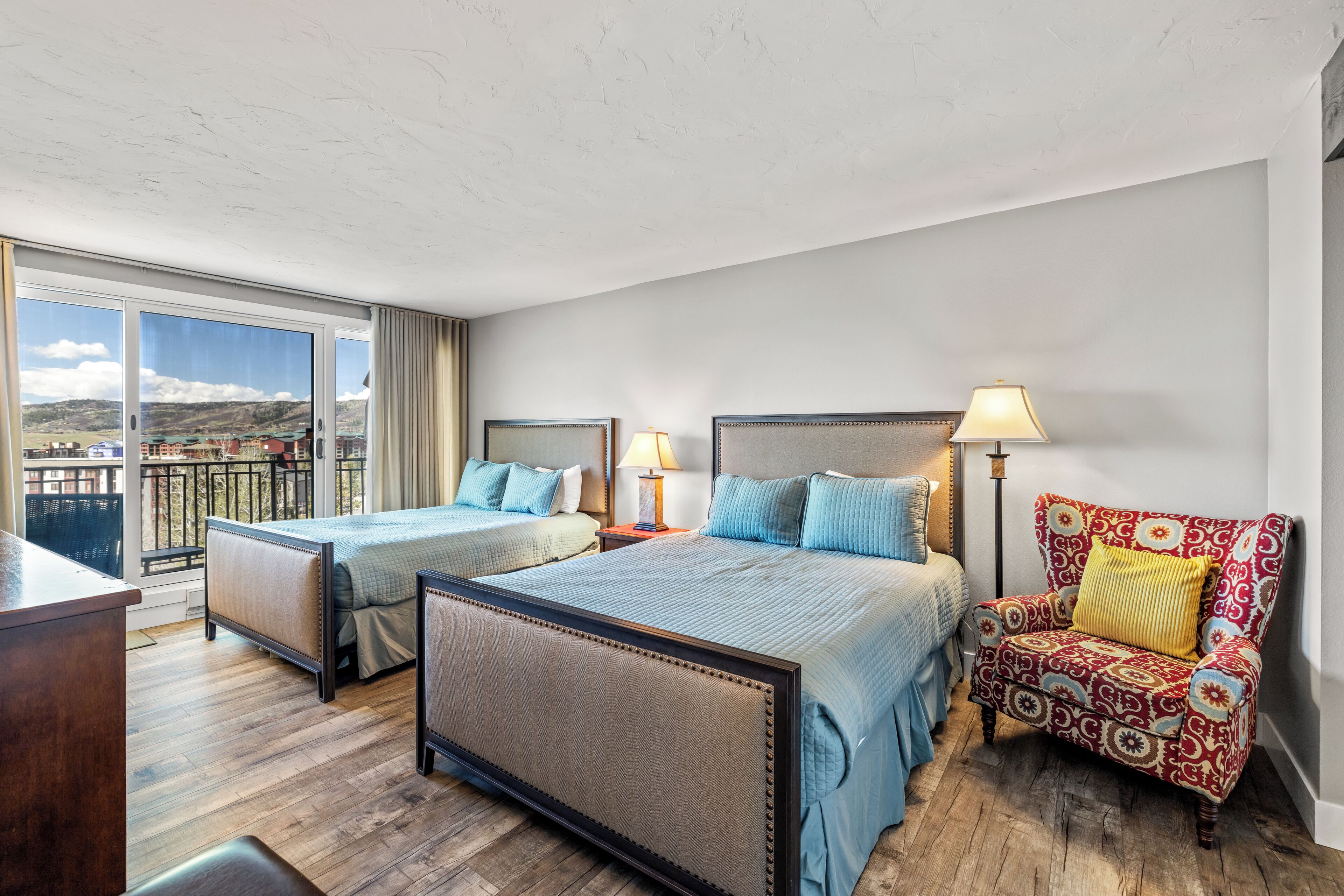A hotel room with two beds, a chair, a desk, and a view of the outdoors.