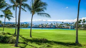A golf course with a pool, palm trees, and a view of a resort with multiple buildings. - Fairway Villas Waikoloa G31 2 Bedroom Villa (Waikoloa)