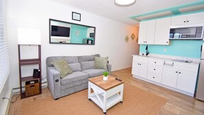 Un espacio habitable funcional con un sofá gris, una pequeña mesa de madera, una zona de cocina con armarios blancos y un televisor montado.