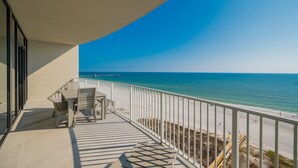 A balcony with a table and chairs, overlooking a beach with a pier in the distance.
