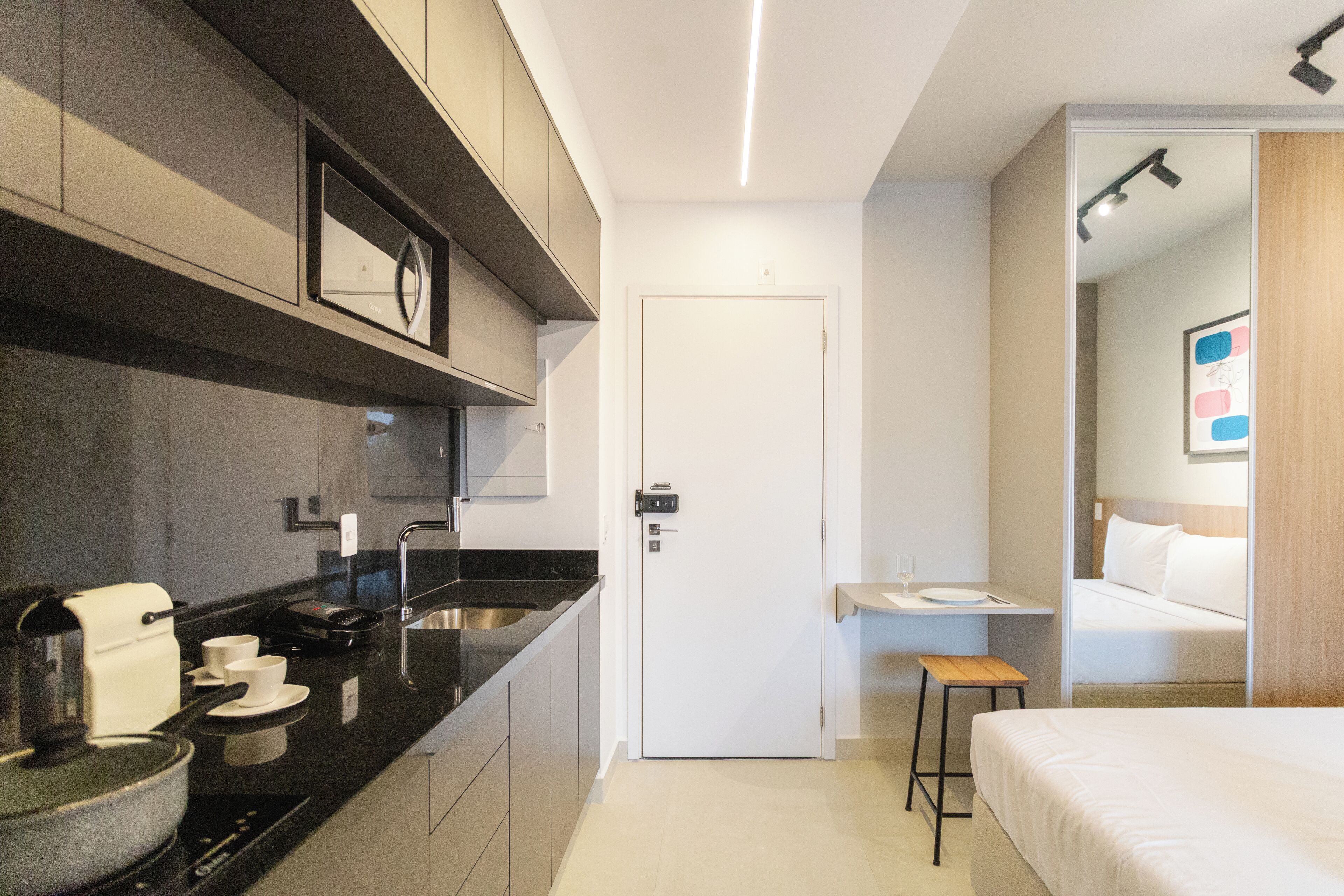 A modern hotel room with a kitchenette, a bed, a small desk, and a bathroom visible through an open door.