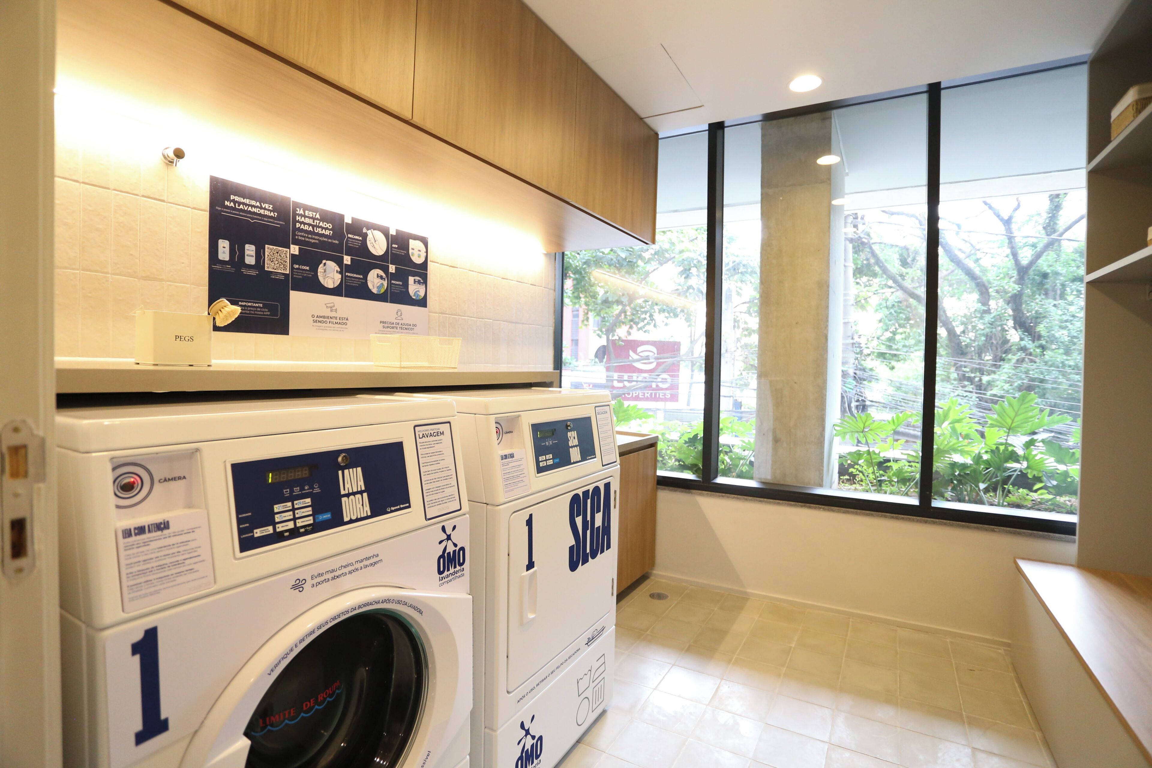 A laundry area with two washing machines, a control panel, and a window with a view of greenery outside.