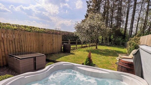 A hot tub in a backyard with a wooden fence, a wooden deck, and a grassy area with trees.