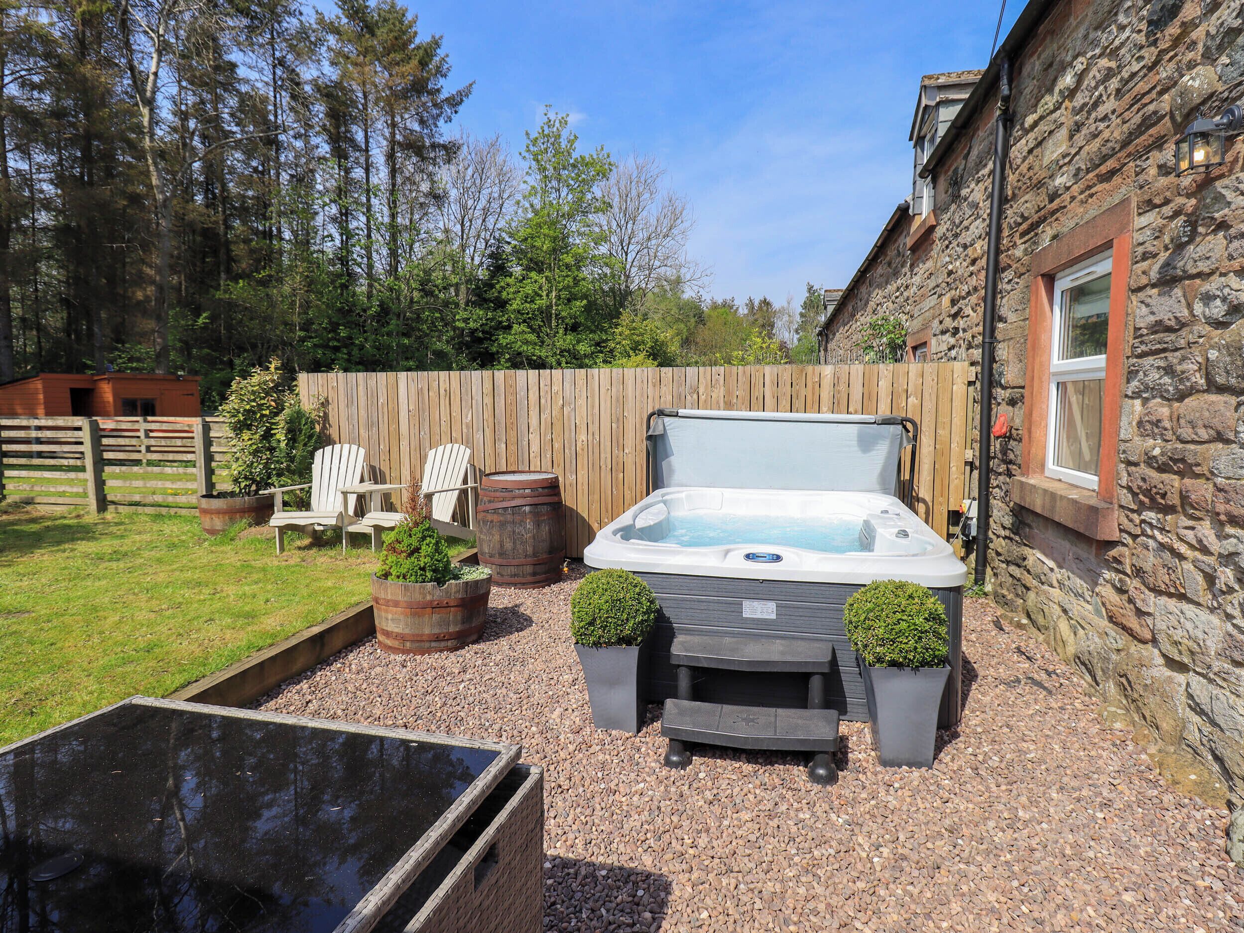 A hot tub in a backyard with a wooden fence, a wooden deck, and a stone building.