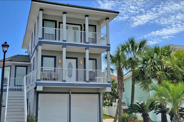 A two-story building with a balcony on the upper floor, a garage door on the lower floor, and palm trees in the background.