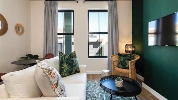A modern living room with a white sofa, a round coffee table, a wicker chair, and a TV mounted on a green wall.