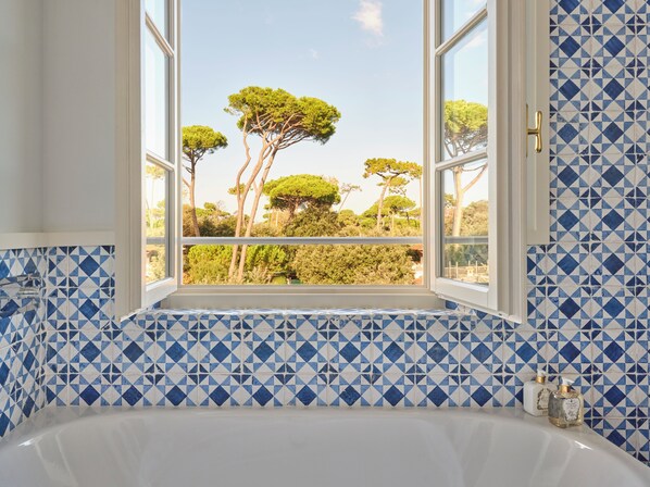 A bathroom with a bathtub, blue and white patterned tiles, and a window with a view of trees. - Pensione America (Forte dei Marmi)