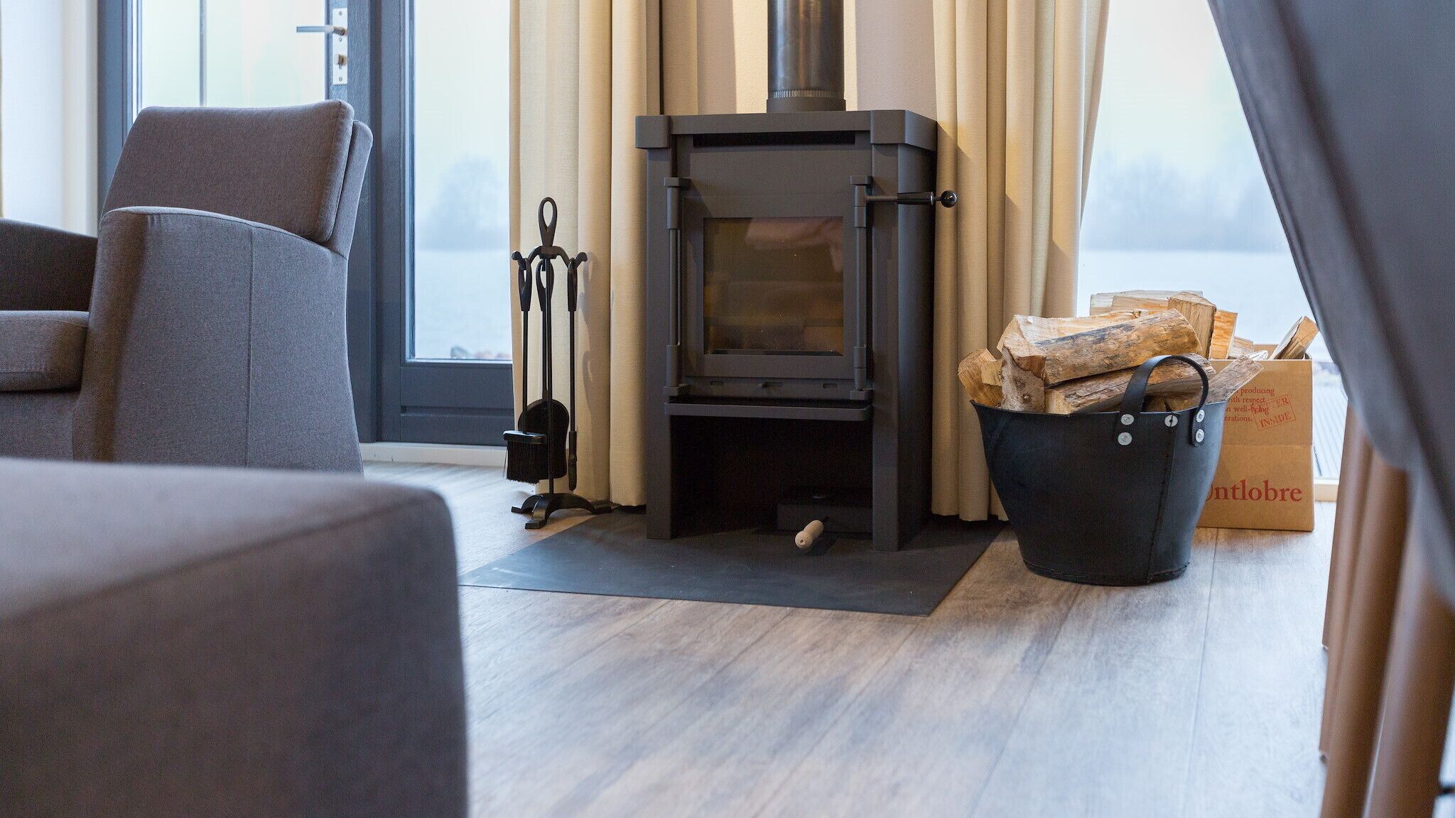 A modern living room with a wood-burning stove, a comfortable grey sofa, and a view through a large window.
