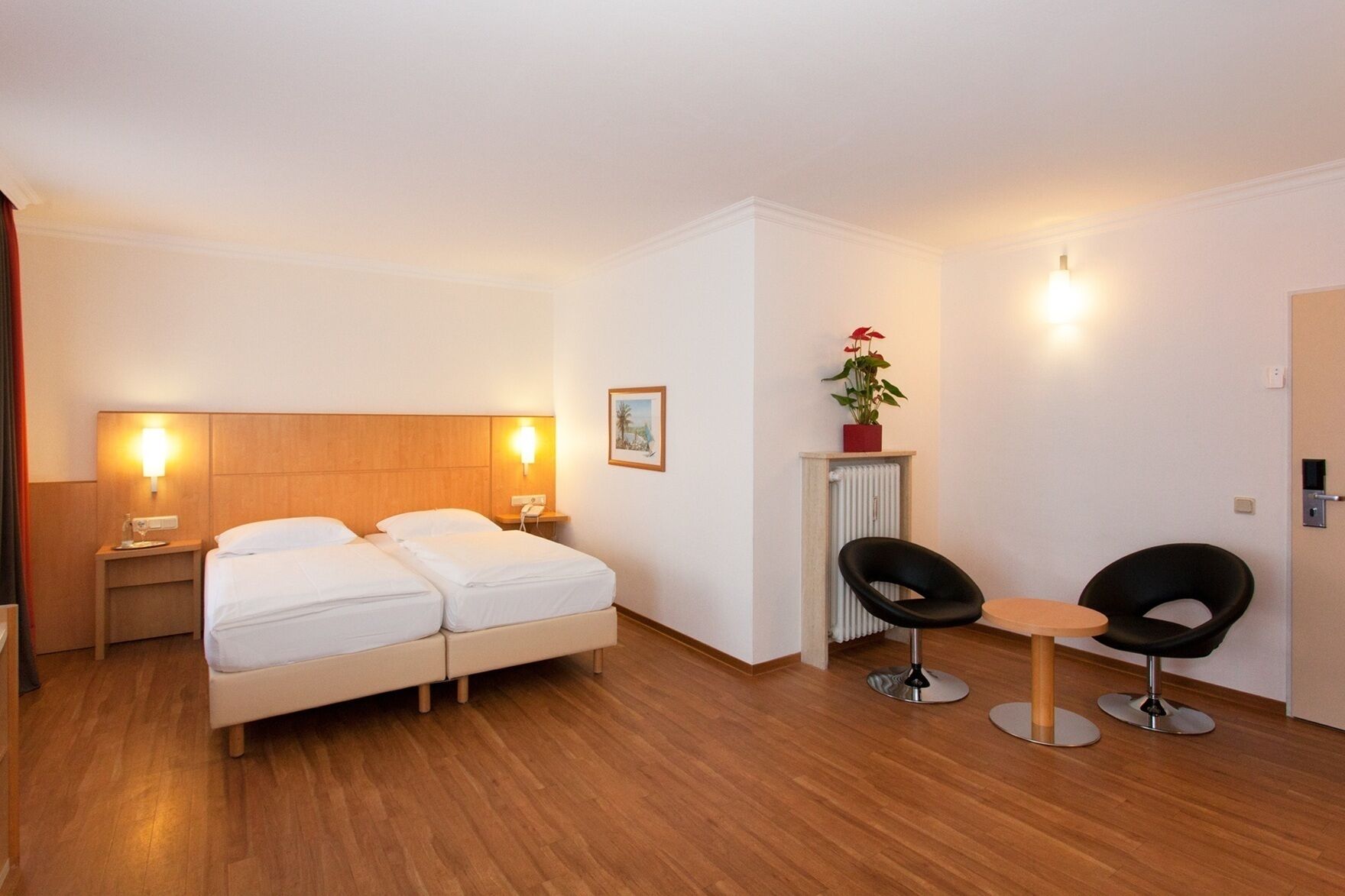 Ein Hotelzimmer mit Doppelbett, Nachttischen mit Lampen, einem Heizkörper, einem kleinen Tisch mit zwei Stühlen und einer Zimmerpflanze.