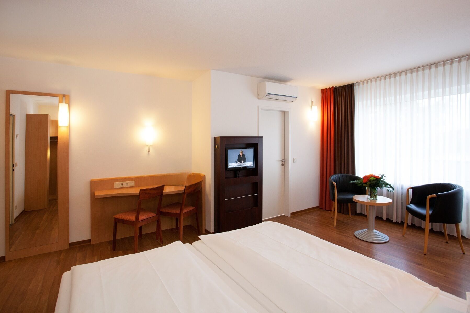 A hotel room with a bed, a desk with two chairs, a television, a small table with a vase of flowers, and a window with curtains.