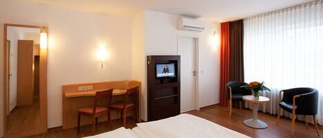 Habitación de hotel con cama, escritorio y dos sillas, televisor, una mesita con un florero y ventana con cortinas.
