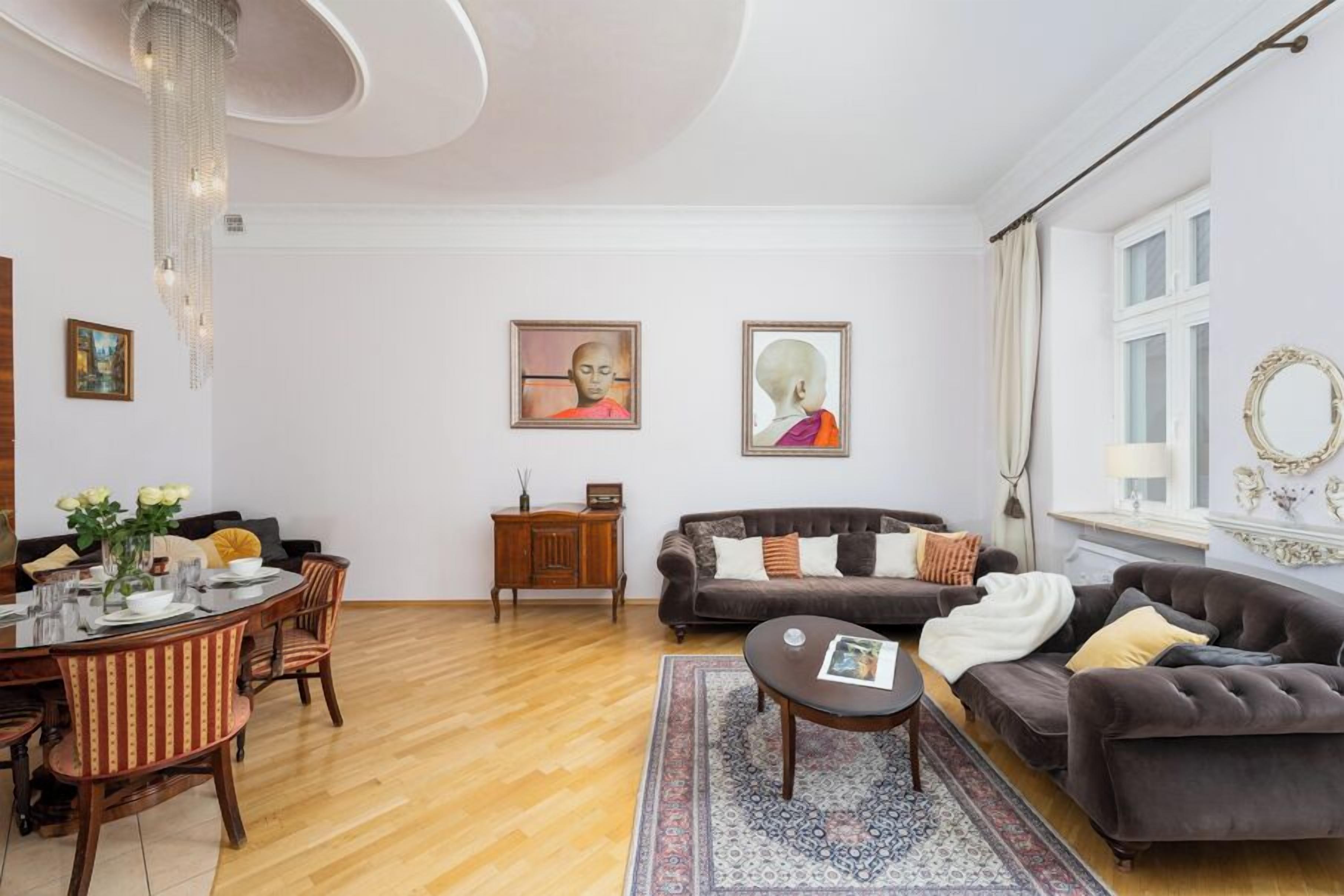 Una sala de estar con comedor, que incluye una araña, un conjunto de sofá, una mesa de centro y obras de arte en las paredes.