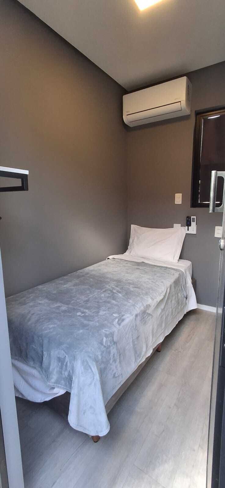 Quarto pequeno, com cama de solteiro, edredom cinza, ar condicionado fixado na parede e mesa de cabeceira com luminária.