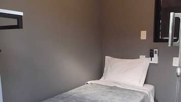 Quarto pequeno, com cama de solteiro, edredom cinza, ar condicionado fixado na parede e mesa de cabeceira com luminária.