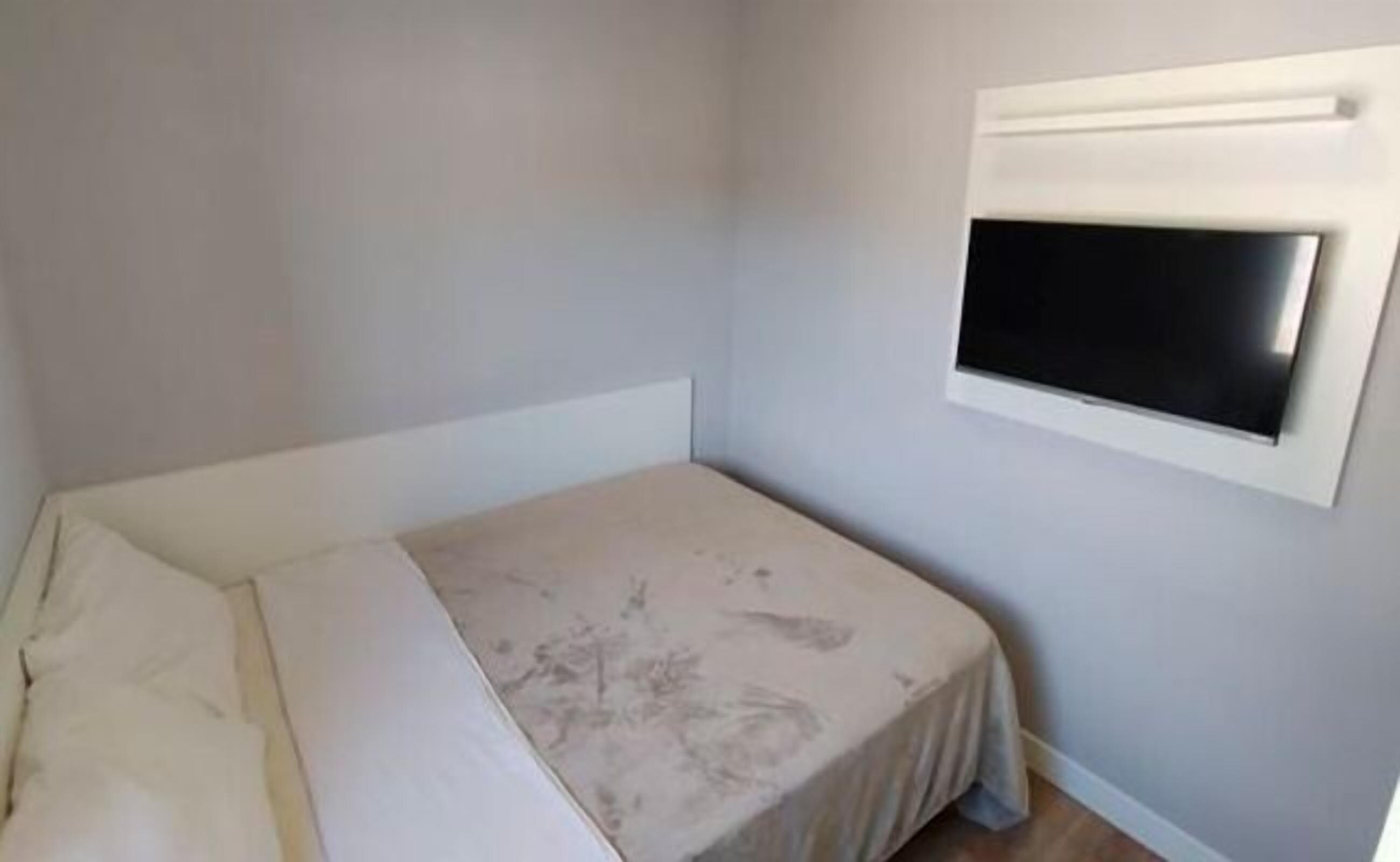 Een kleine kamer met een enkel bed, een wandtelevisie en een effen wand.