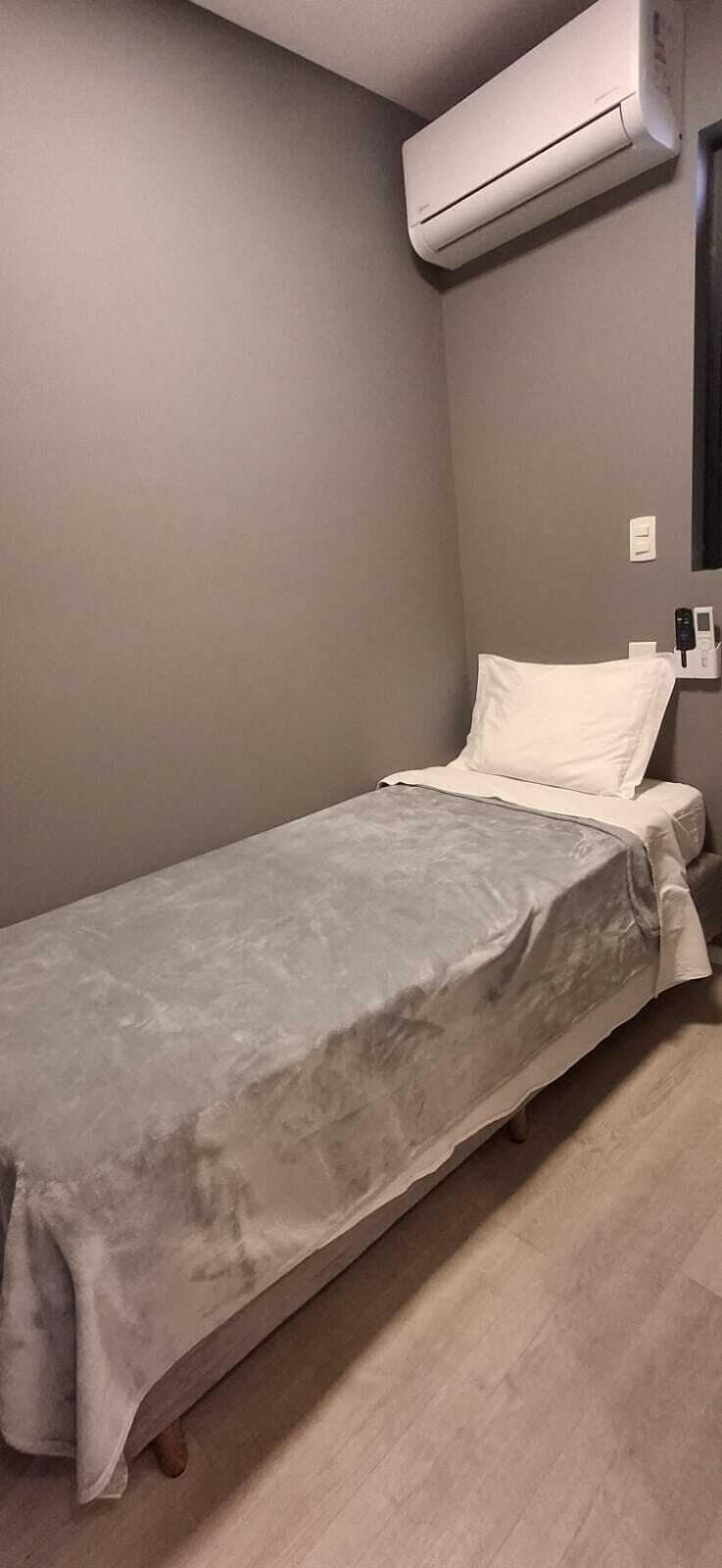 Un lit simple avec une couvre-lit grise, dans une pièce dont le mur est également gris et où un appareil de climatisation est fixé au-dessus.