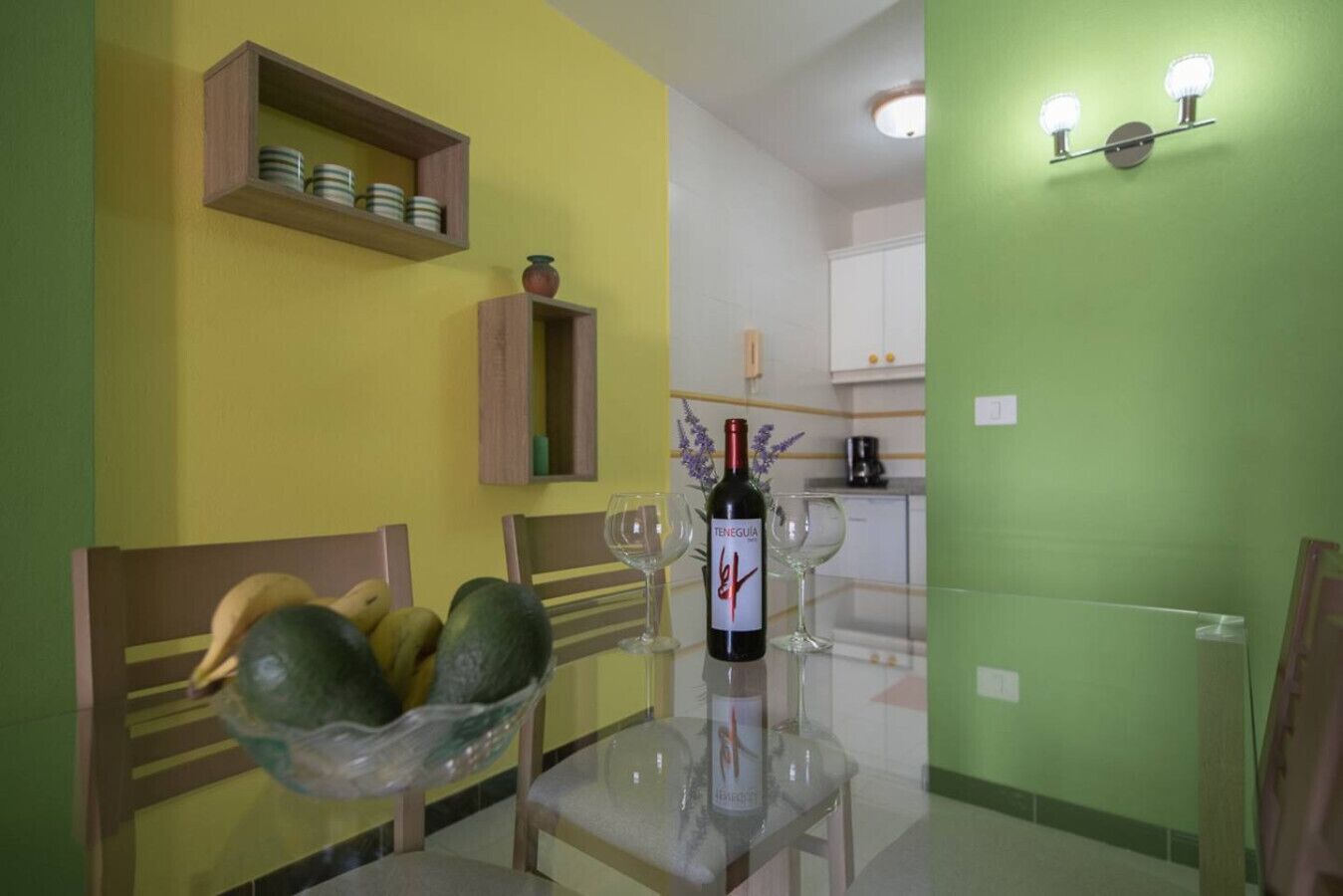 Un comedor con una mesa de vidrio, sillas, un cuenco de fruta y una botella de vino.