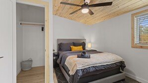 Un dormitorio con cama, ventilador de techo, ventana con persianas, mesita de noche y cesta de ropa.