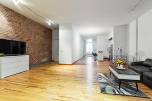 A modern living room with a brick wall, wooden flooring, a flat-screen TV, a white entertainment unit, a black sofa, a coffee table, and a dining area with a glass table and chairs.