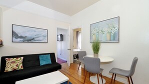 Una sala de estar moderna con un sofá negro, una mesa de centro blanca y sillas grises alrededor de una pequeña mesa blanca. Hay dos cuadros enmarcados en las paredes y un televisor montado en la pared.
