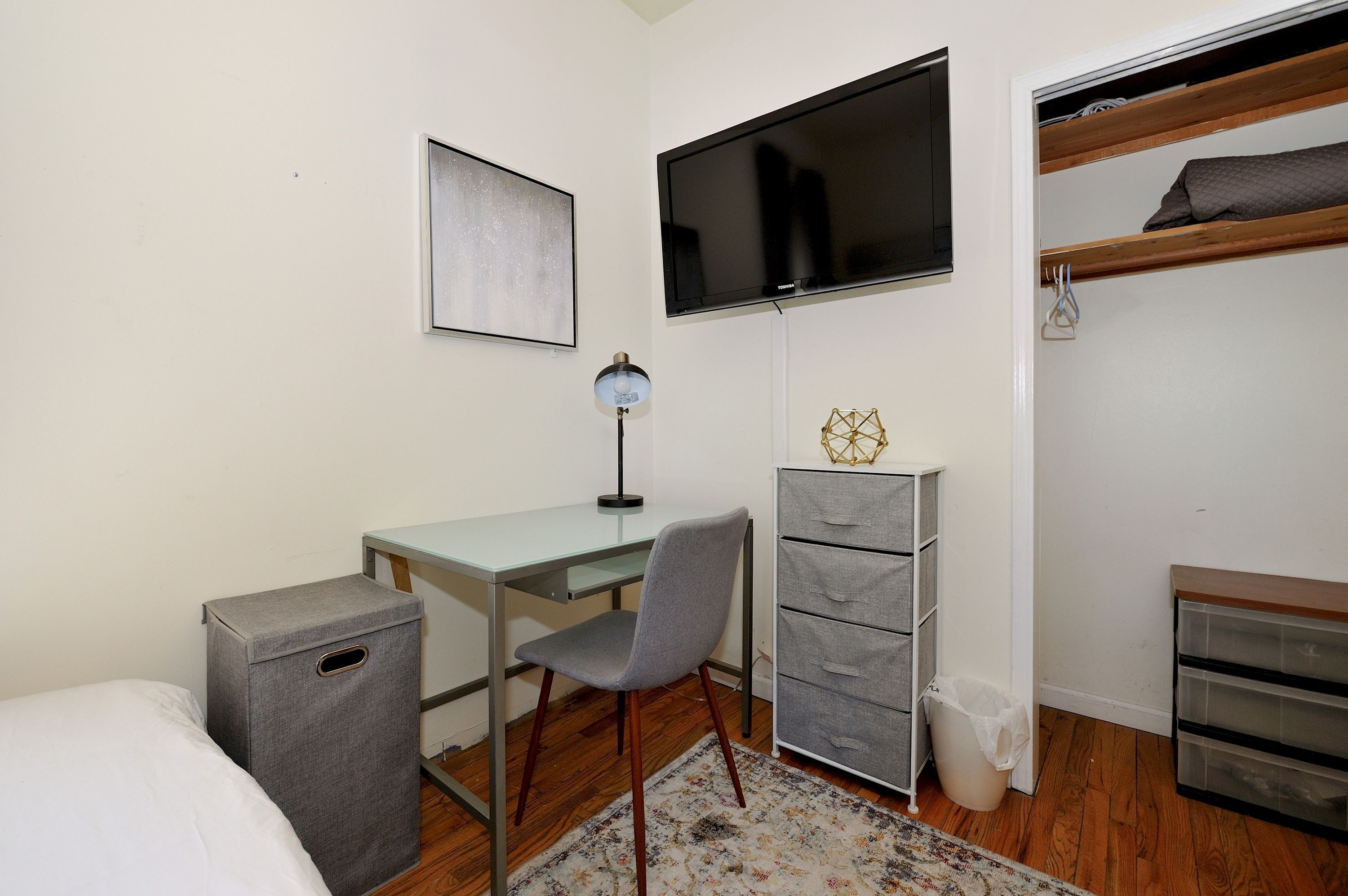 A small, neatly arranged bedroom with a bed, a desk, a chair, a TV, a wardrobe, and a trash bin.
