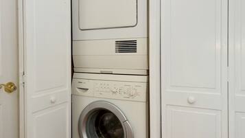A compact, white laundry room with a front-loading washing machine and dryer, a small shelf with cleaning supplies, and a wooden floor.