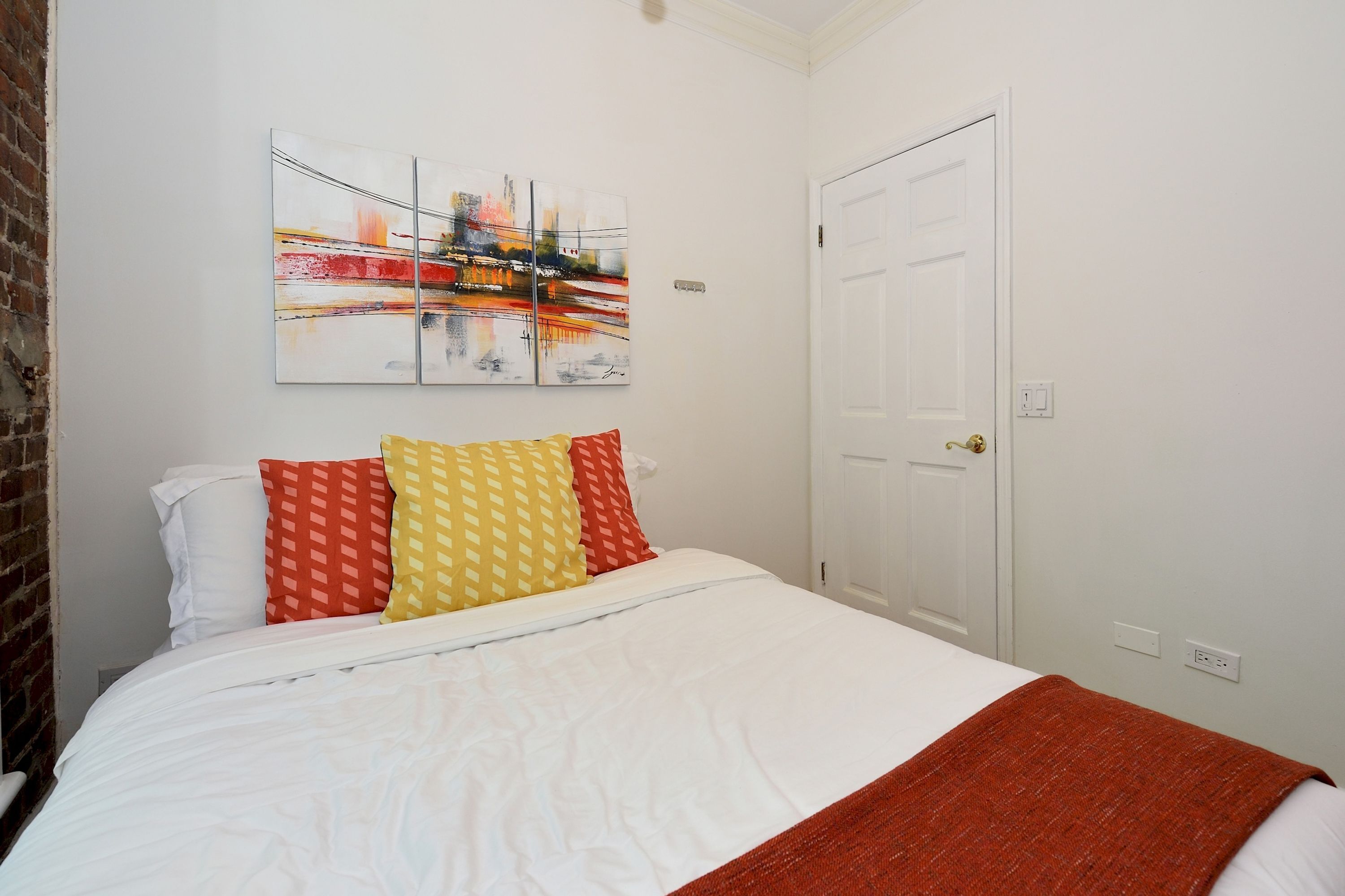 Quarto de hotel com cama branca, almofadas coloridas, manta vermelha e pintura de paisagem urbana na parede.