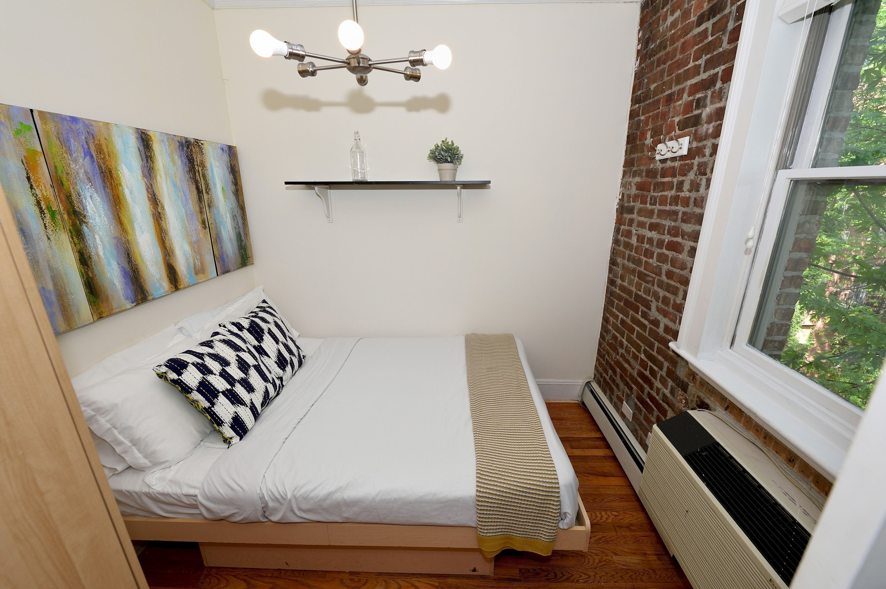 Un dormitorio pequeño y acogedor con cama, lámpara colgante, estante en la pared con una planta y ventana con vistas a follaje.