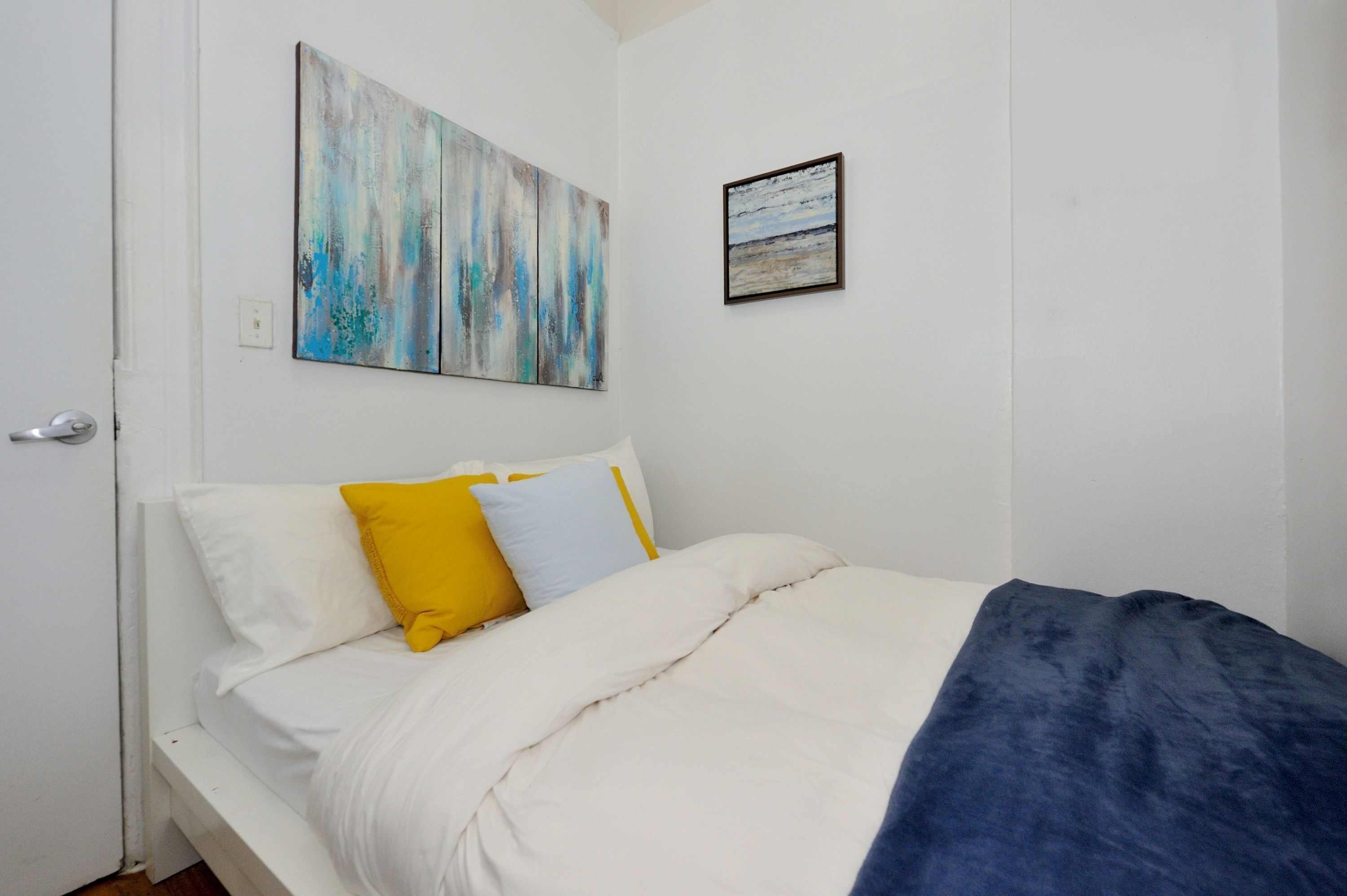 A bedroom with a bed, white walls, a door, and two paintings.