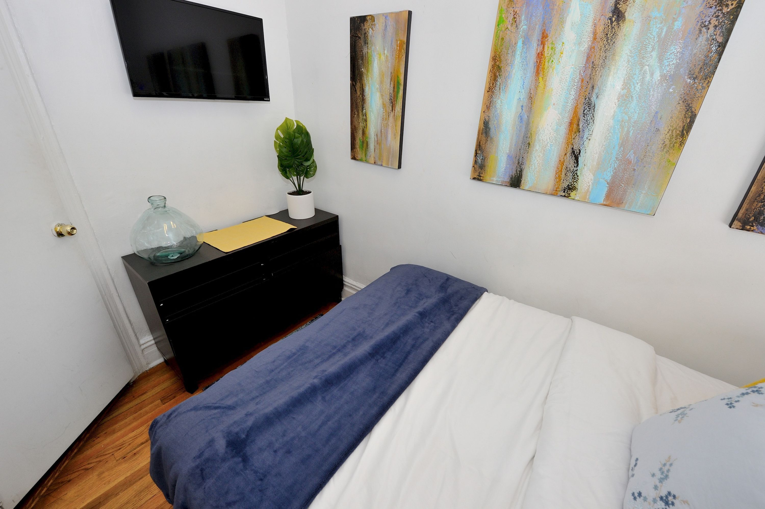Ein Schlafzimmer mit Bett, Schminktisch, Zimmerpflanze und abstrakten Wandbildern.
