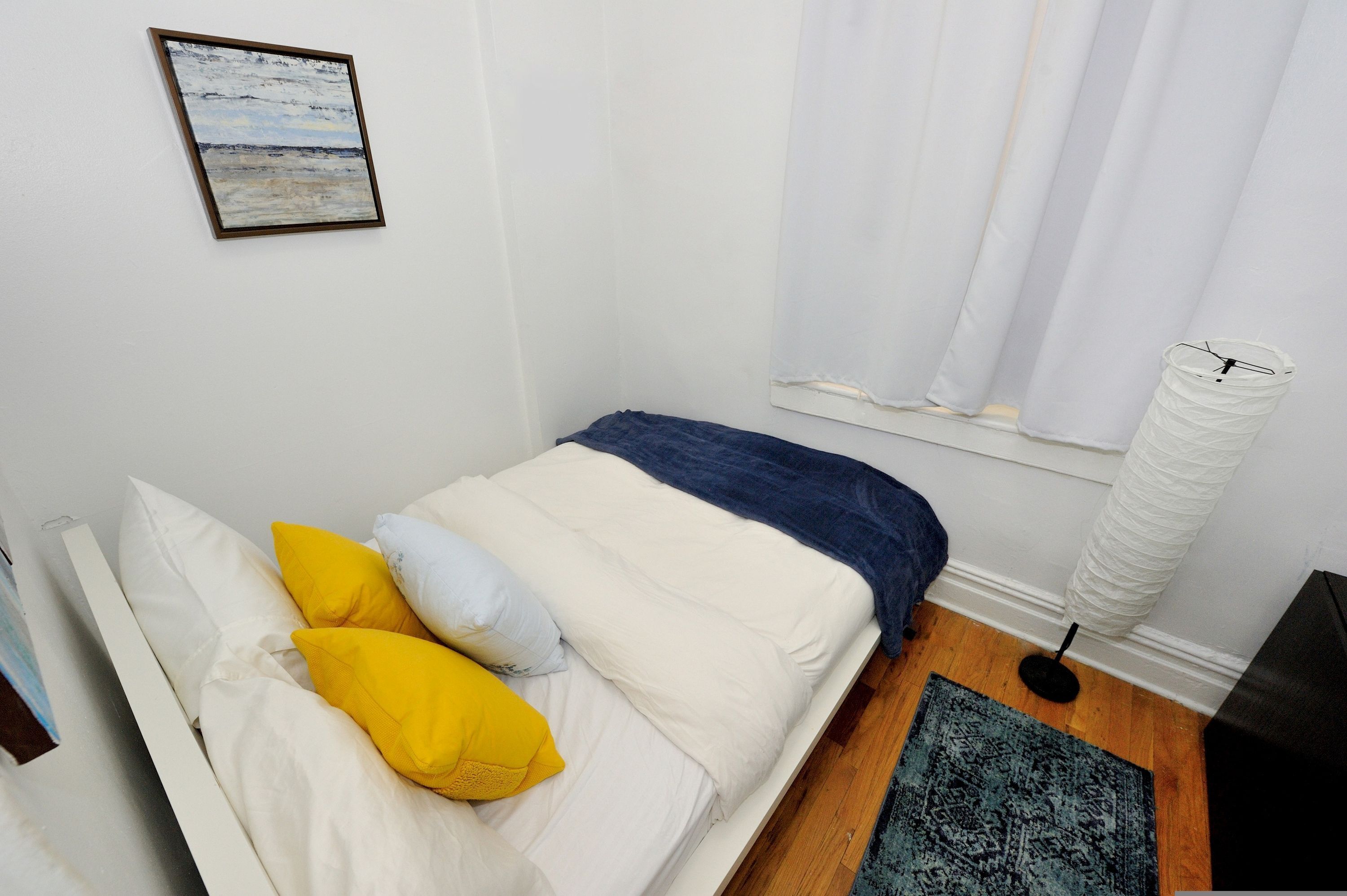 Una cama bien hecha con almohadas blancas y amarillas, una manta azul y un cabecero blanco. Encima de la cama, un cuadro enmarcado cuelga de la pared.