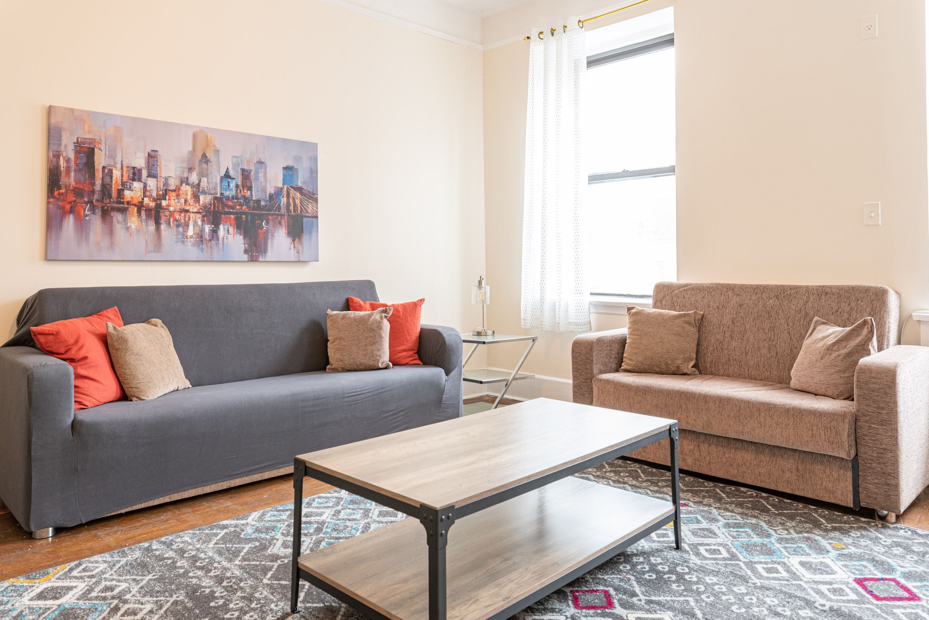 Una sala de estar con un sofá gris, un sillón color beige y una mesa de centro de madera. En la pared, un cuadro con un paisaje urbano, y se ve una ventana con cortinas.