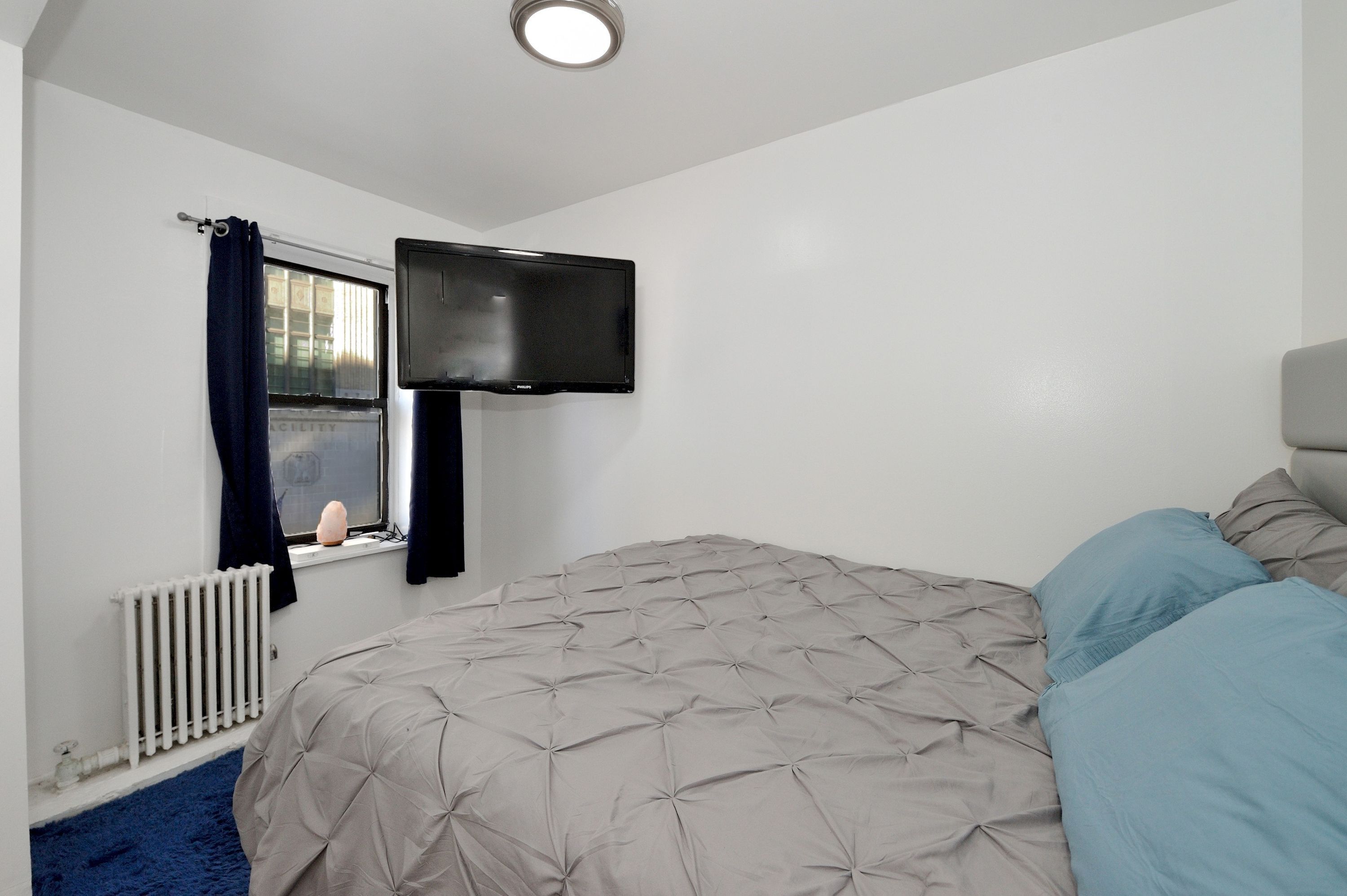 A bedroom with a bed, a flat-screen TV mounted on the wall, a window with curtains, and a radiator.